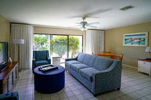 A living room with a sofa, a round ottoman, a television, and a ceiling fan. - Ventura at Boca Raton (Boca Raton)