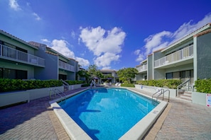 A rectangular pool with a tiled edge, surrounded by a brick walkway, and adjacent to a building with balconies and a covered seating area. - Ventura at Boca Raton (Boca Raton)