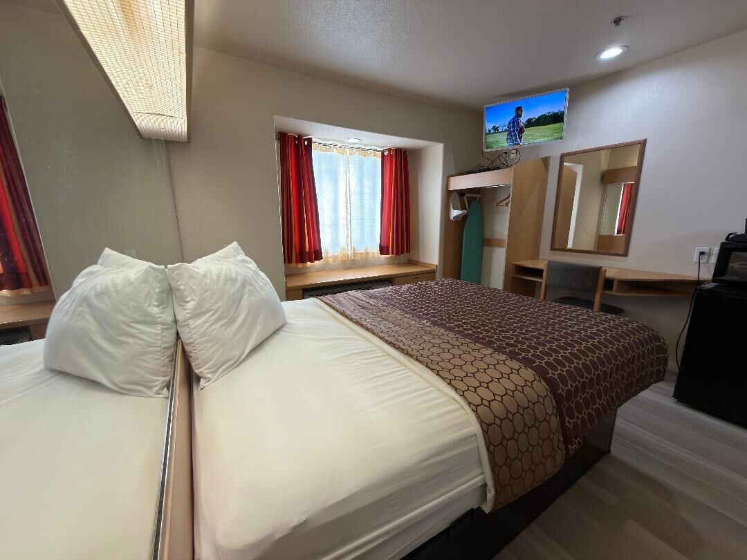 Una habitación de hotel con cama, escritorio, televisión y ventana con cortinas.