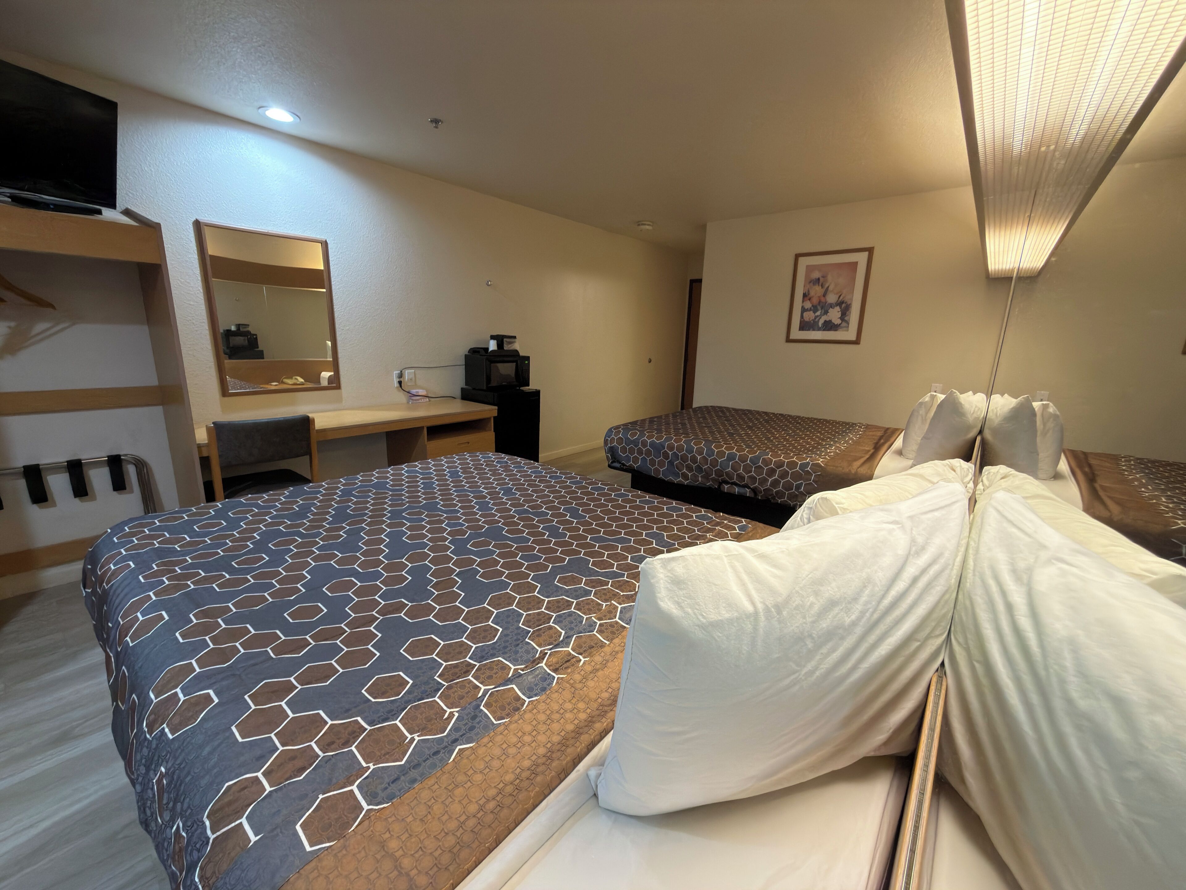 Habitación de hotel con una cama grande, otra más pequeña, un escritorio con una silla, un espejo, un televisor y un cuadro en la pared.