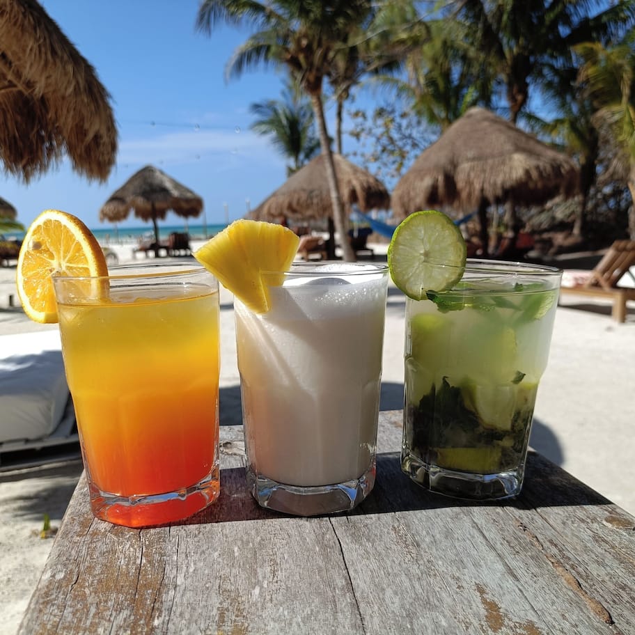 Three drinks on a wooden table with a beach and palm trees in the background.