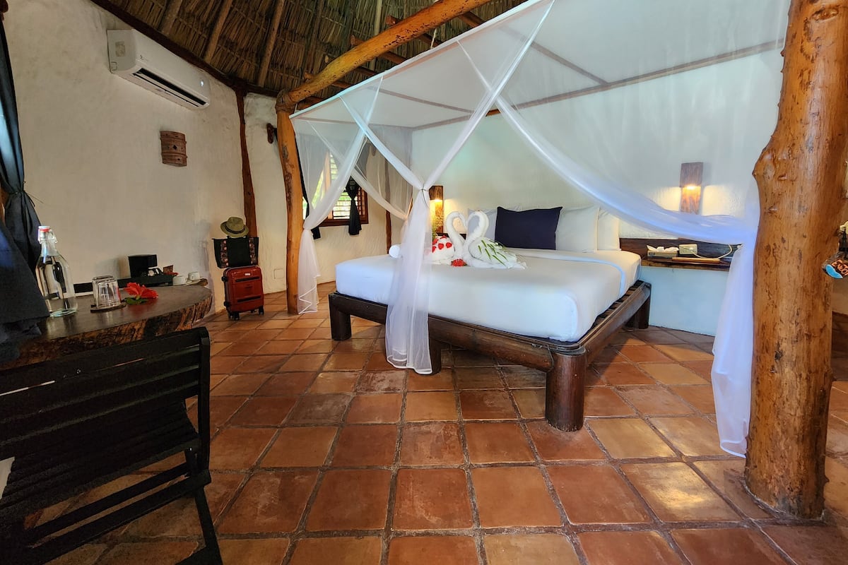 A bedroom with a bed under a canopy, a wooden desk, a chair, a nightstand, and a small table with a lamp.
