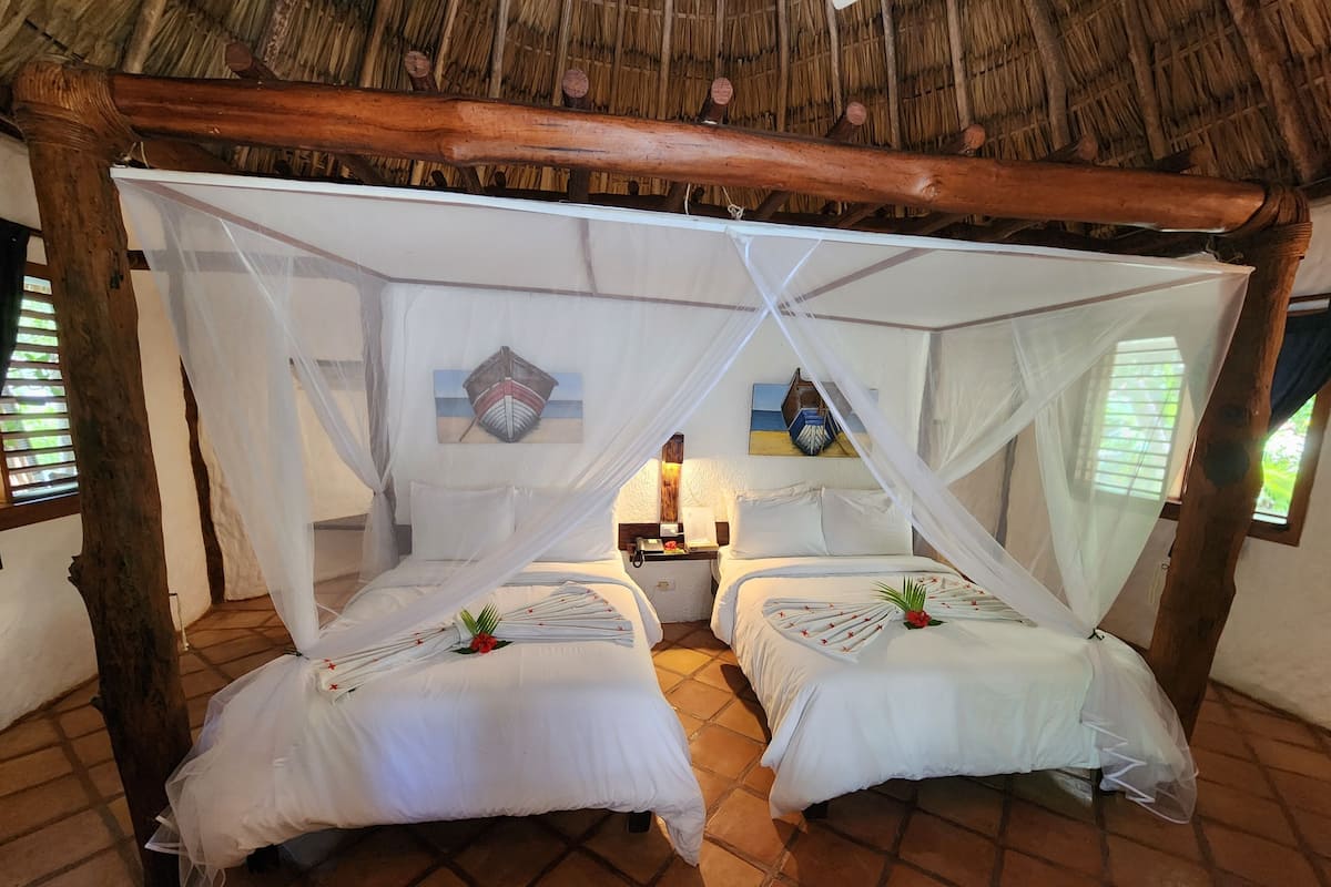 A room with two beds under a thatched roof, white bedding, and a decorative wall hanging.