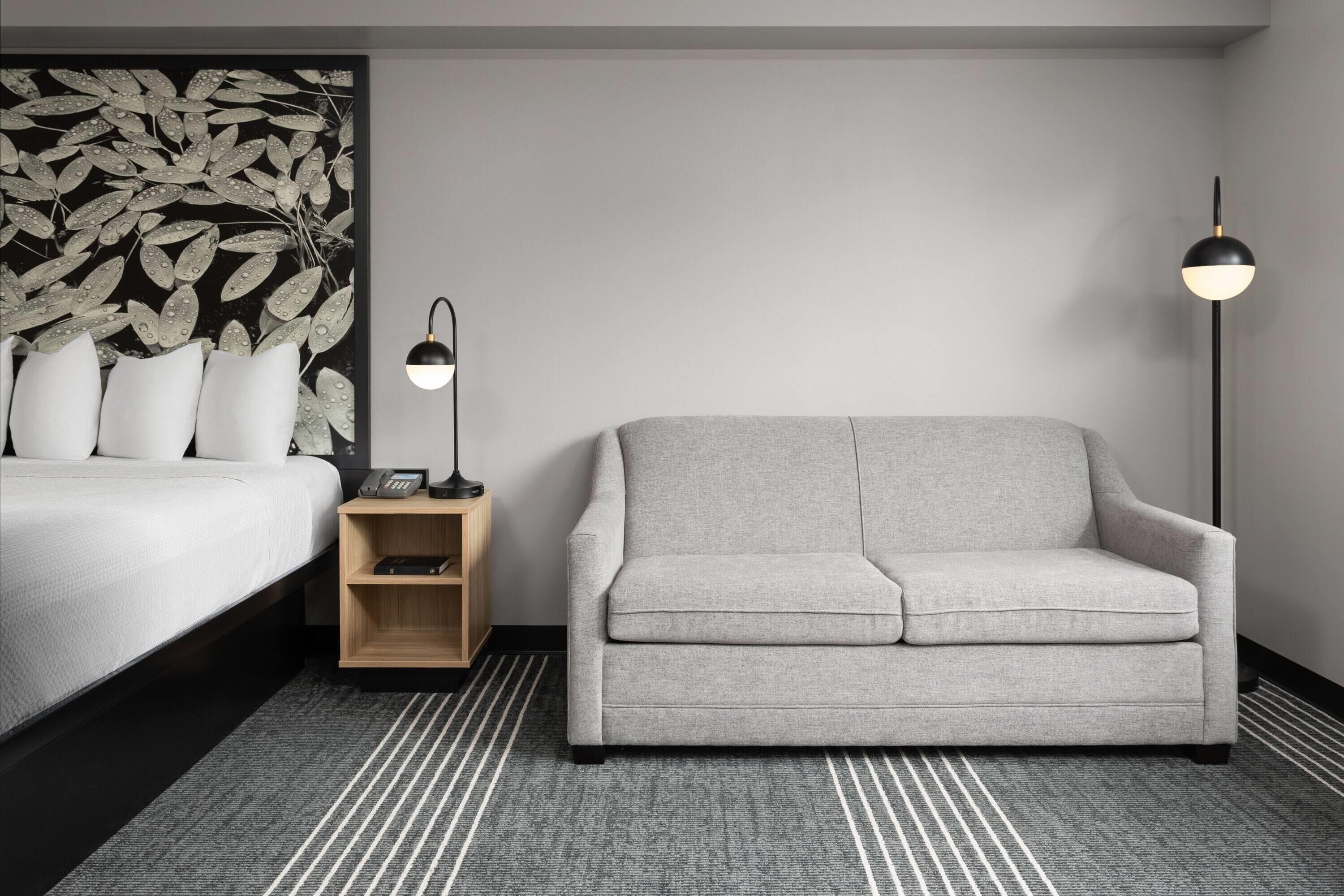 Una habitación de hotel moderna con una cama, un sofá gris, una mesita de noche con lámpara y un panel decorativo en la pared.
