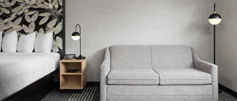 Una habitación de hotel moderna con una cama, un sofá gris, una mesita de noche con lámpara y un panel decorativo en la pared.