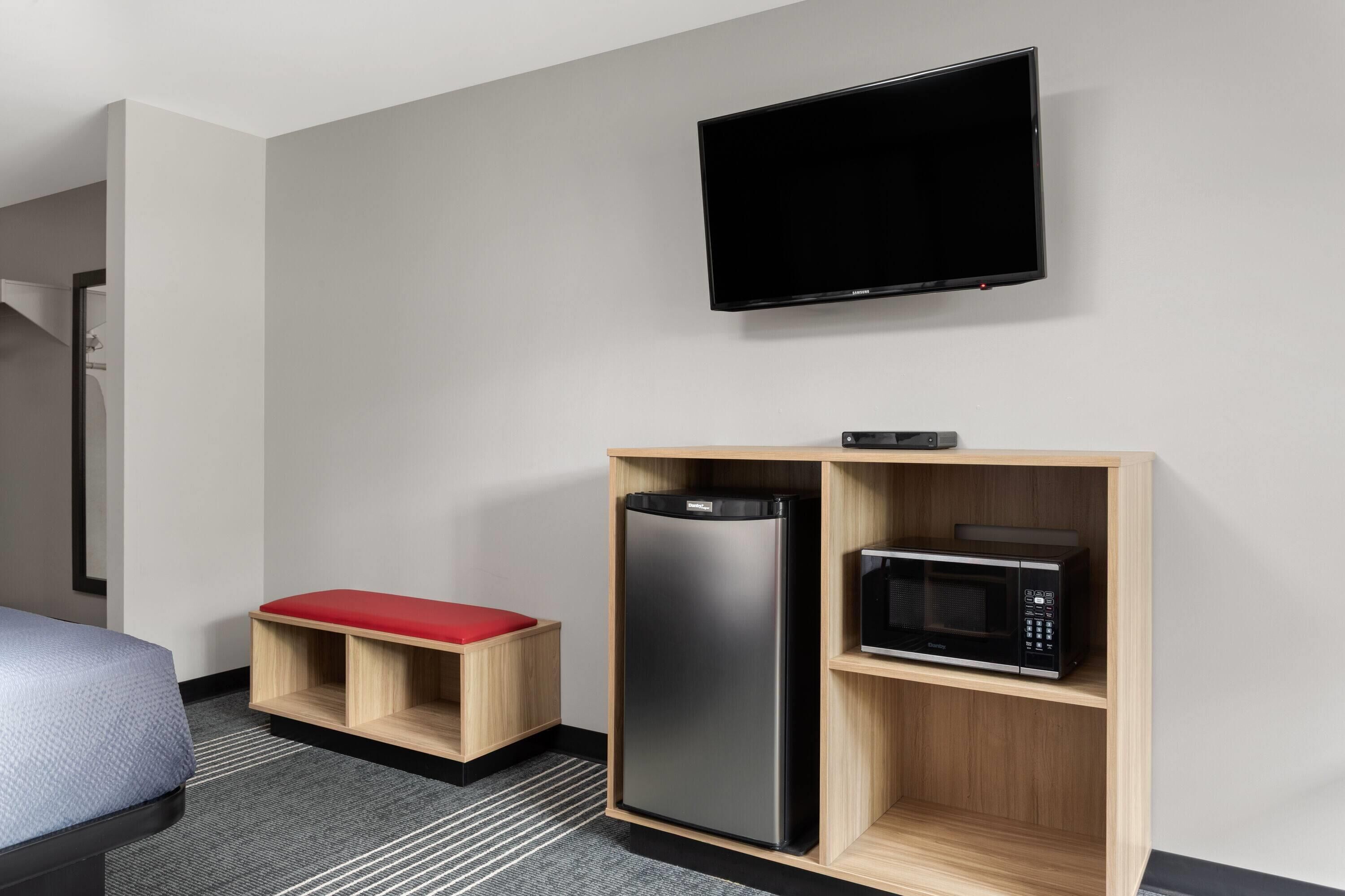Une chambre d’hôtel avec un lit, un téléviseur fixé au mur, un meuble en bois contenant un four à micro-ondes et un banc rouge.