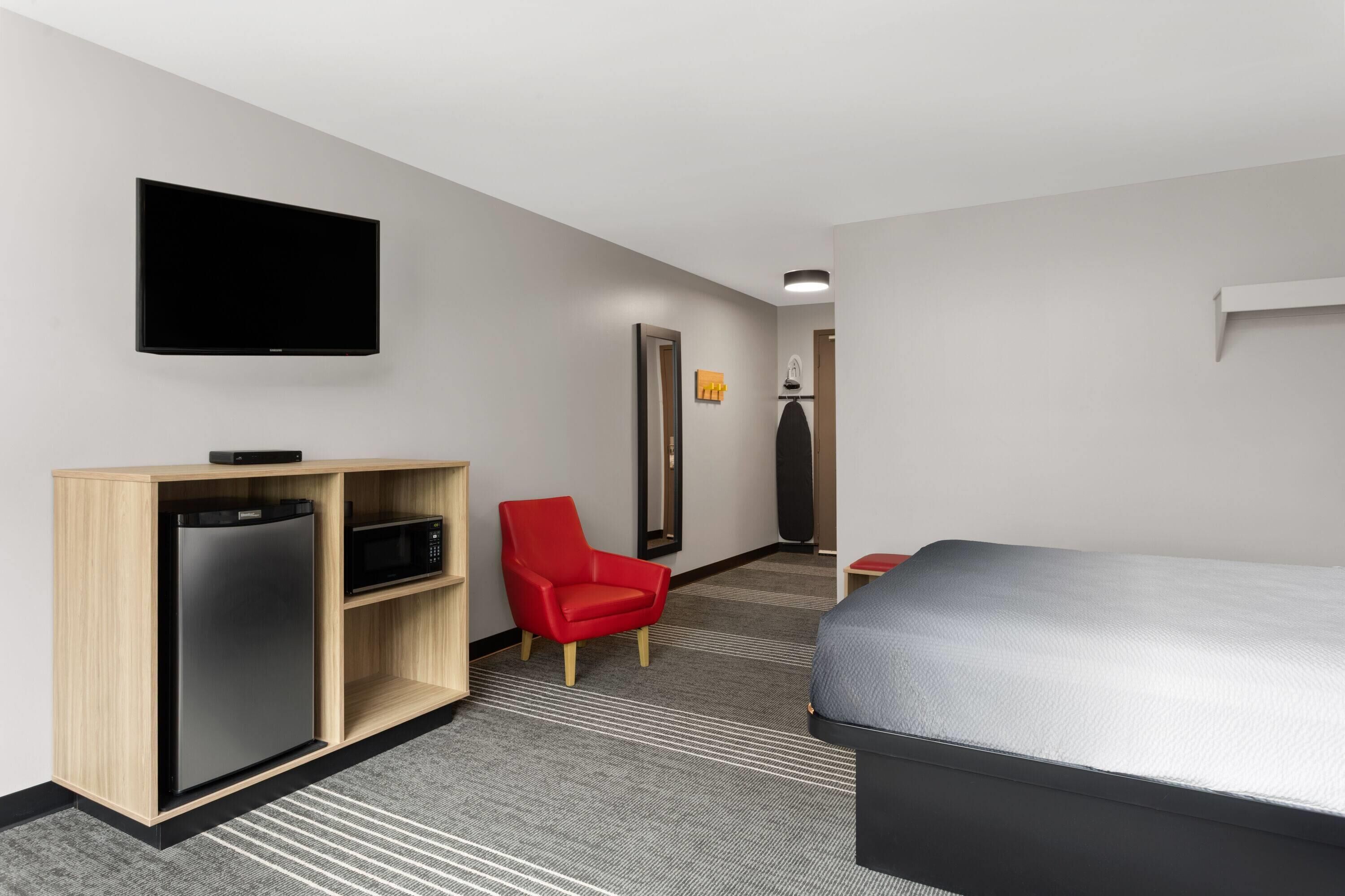 Une chambre d’hôtel avec un lit, un téléviseur fixé au mur, un réfrigérateur, un four à micro-ondes et une chaise rouge.