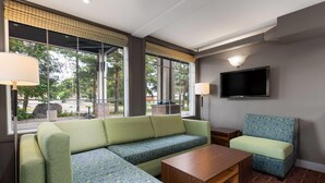 A modern hotel room with a sectional sofa, a matching armchair, a wooden coffee table, and a flat-screen TV mounted on the wall.
