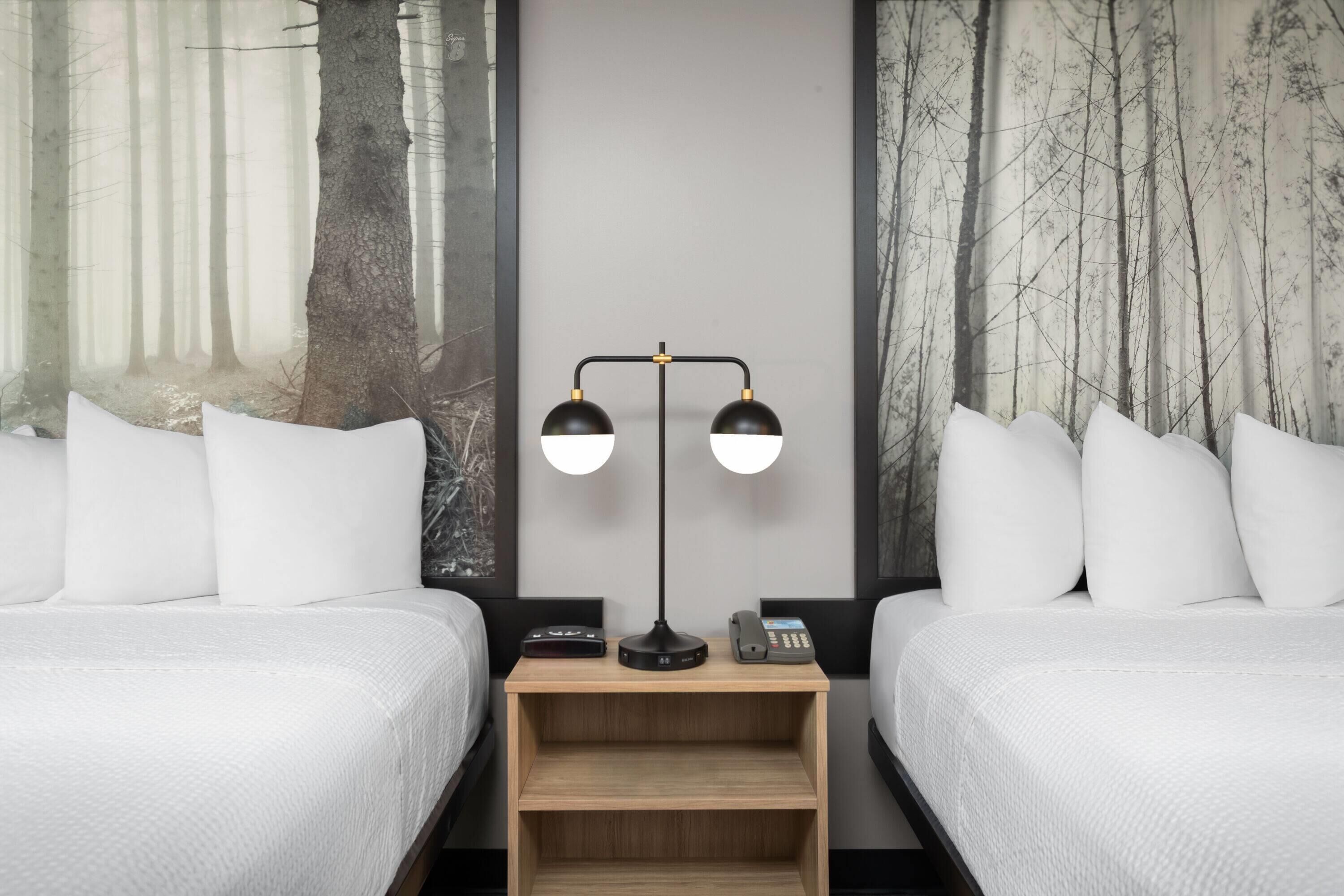 A hotel room with two beds, a nightstand with a lamp, a phone, and a painting of a forest on the wall.