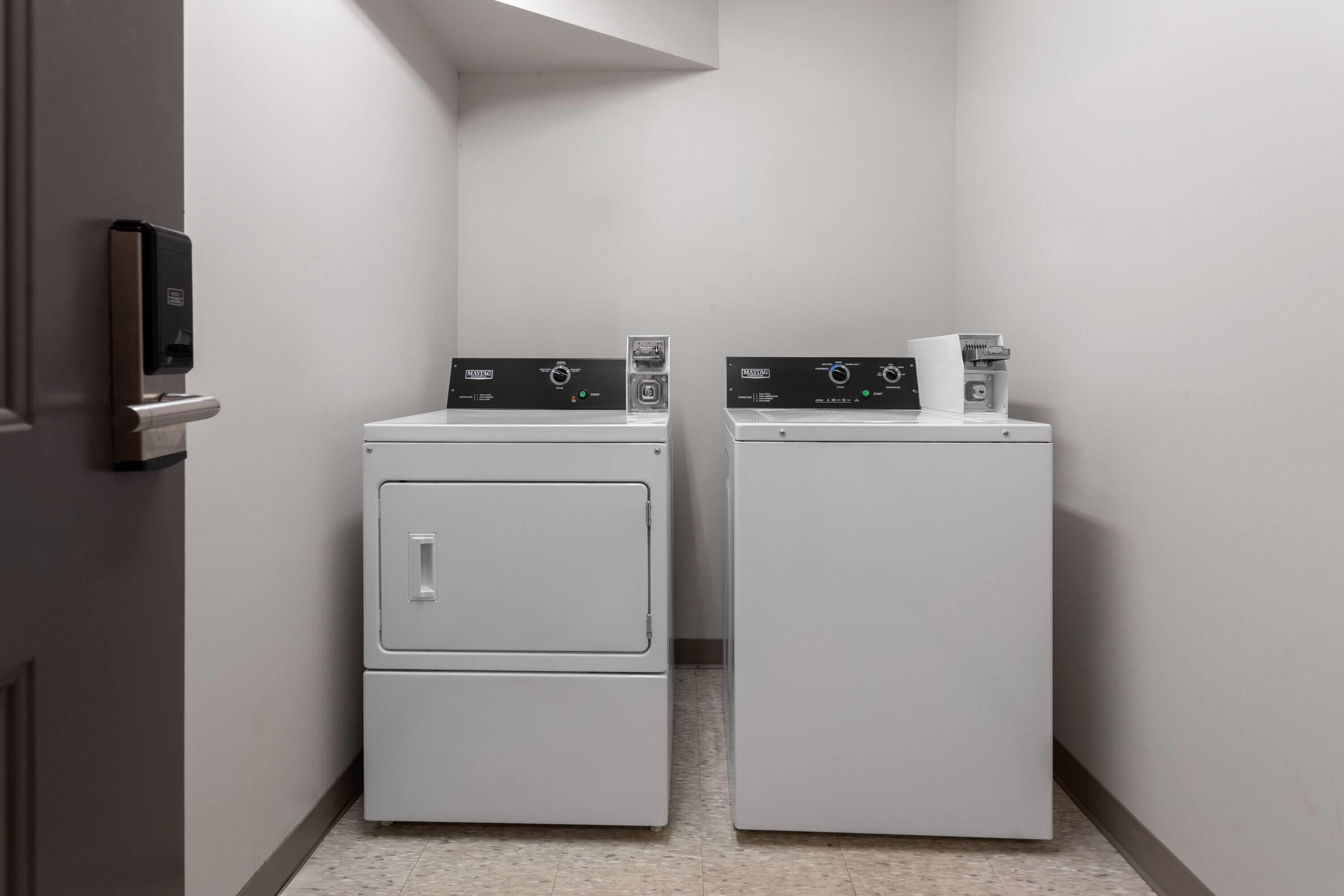A laundry room with two white front-loading washing machines and dryers, a door with a lock, and a small shelf with a paper towel dispenser.