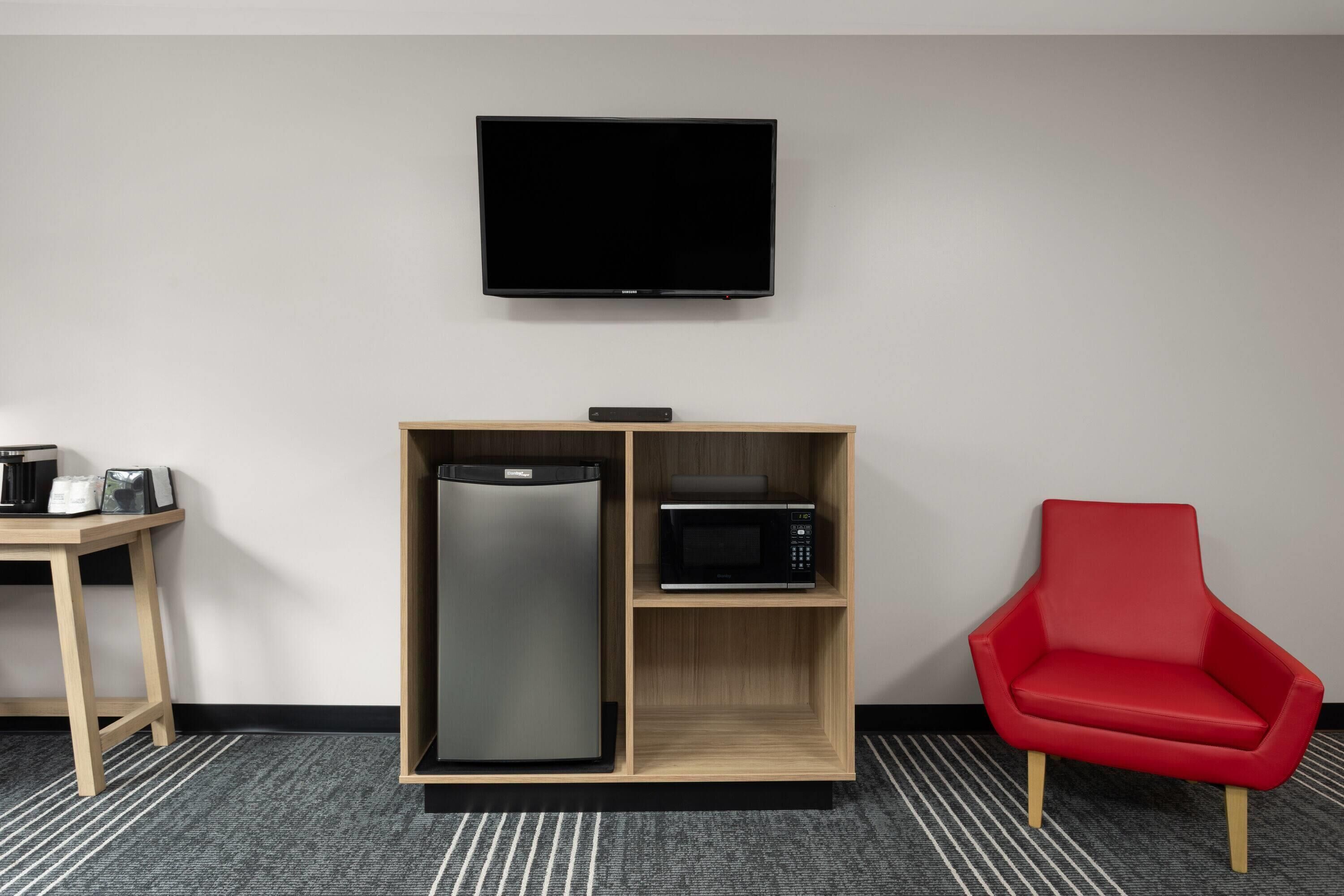 A room with a flat-screen TV mounted on the wall, a wooden cabinet with a refrigerator and microwave, a red chair, and a small table with a coffee maker and a picture frame.