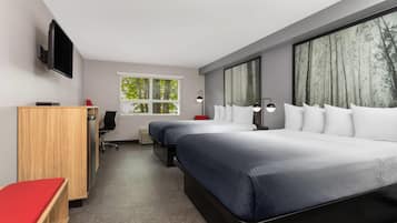 A modern hotel room with two beds, a desk, a chair, a TV, and large windows with a view of trees.