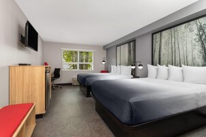 Habitación de hotel moderna con dos camas, un escritorio, una silla, un televisor y ventanas amplias con vista a árboles.