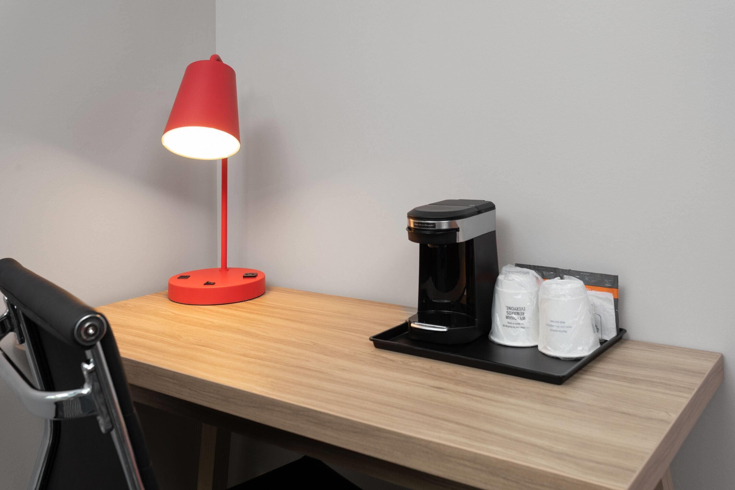 Ein Holztisch mit einer roten Schreibtischlampe, einer Kaffeemaschine und zwei weißen Behältern auf einer Ablagefläche.