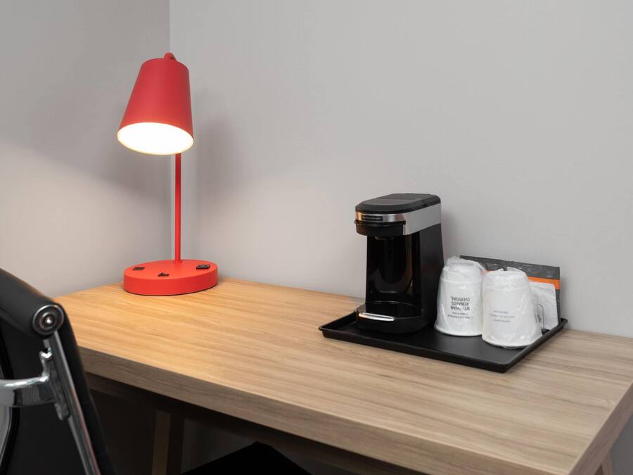 Ein Holztisch mit einer roten Schreibtischlampe, einer Kaffeemaschine und zwei weißen Behältern auf einer Ablagefläche.