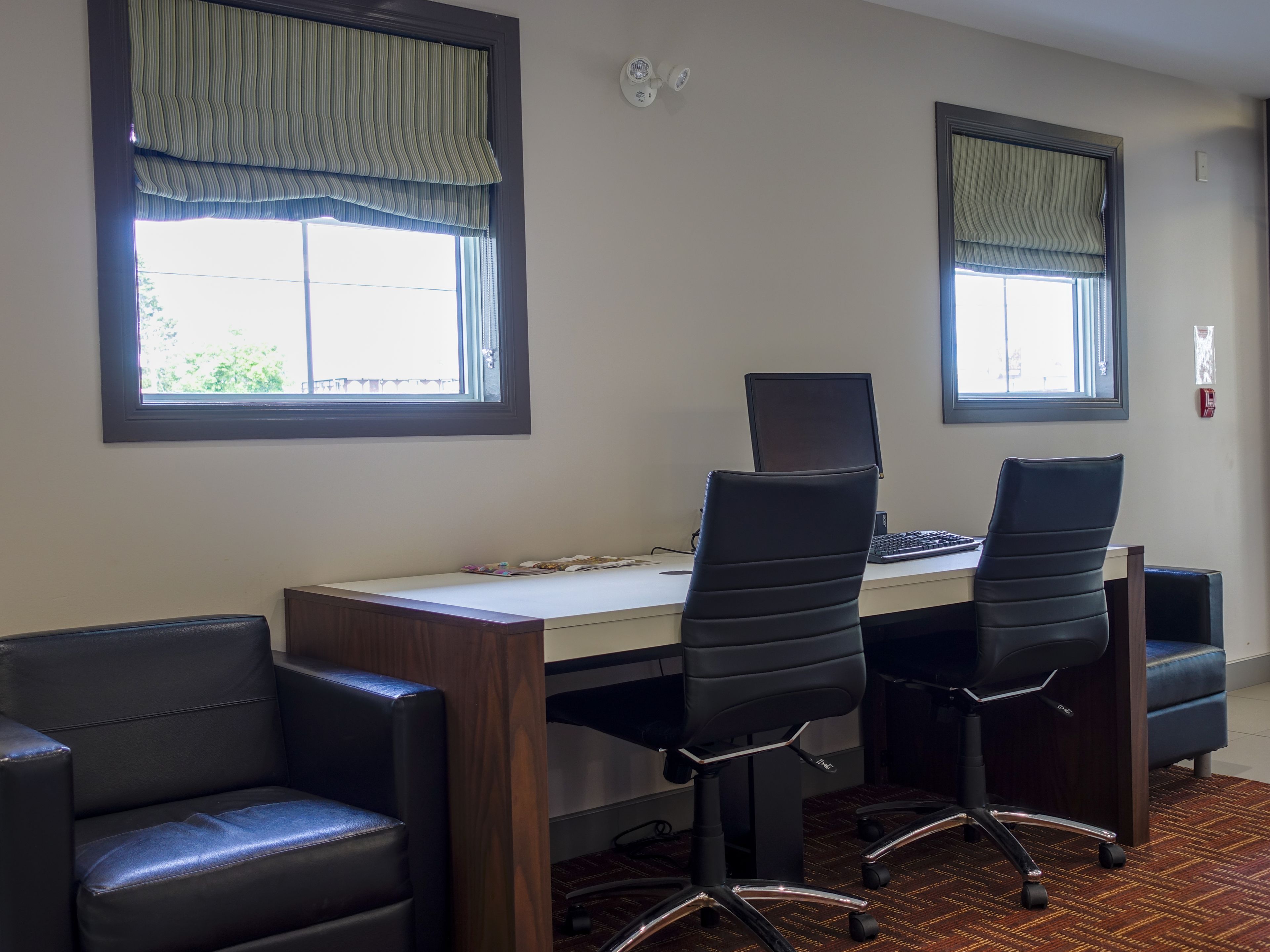 Quarto com escrivaninha, duas cadeiras de escritório pretas, um computador e duas janelas com cortinas verticais.
