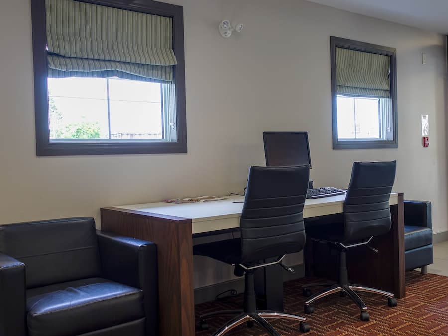 Quarto com escrivaninha, duas cadeiras de escritório pretas, um computador e duas janelas com cortinas verticais.