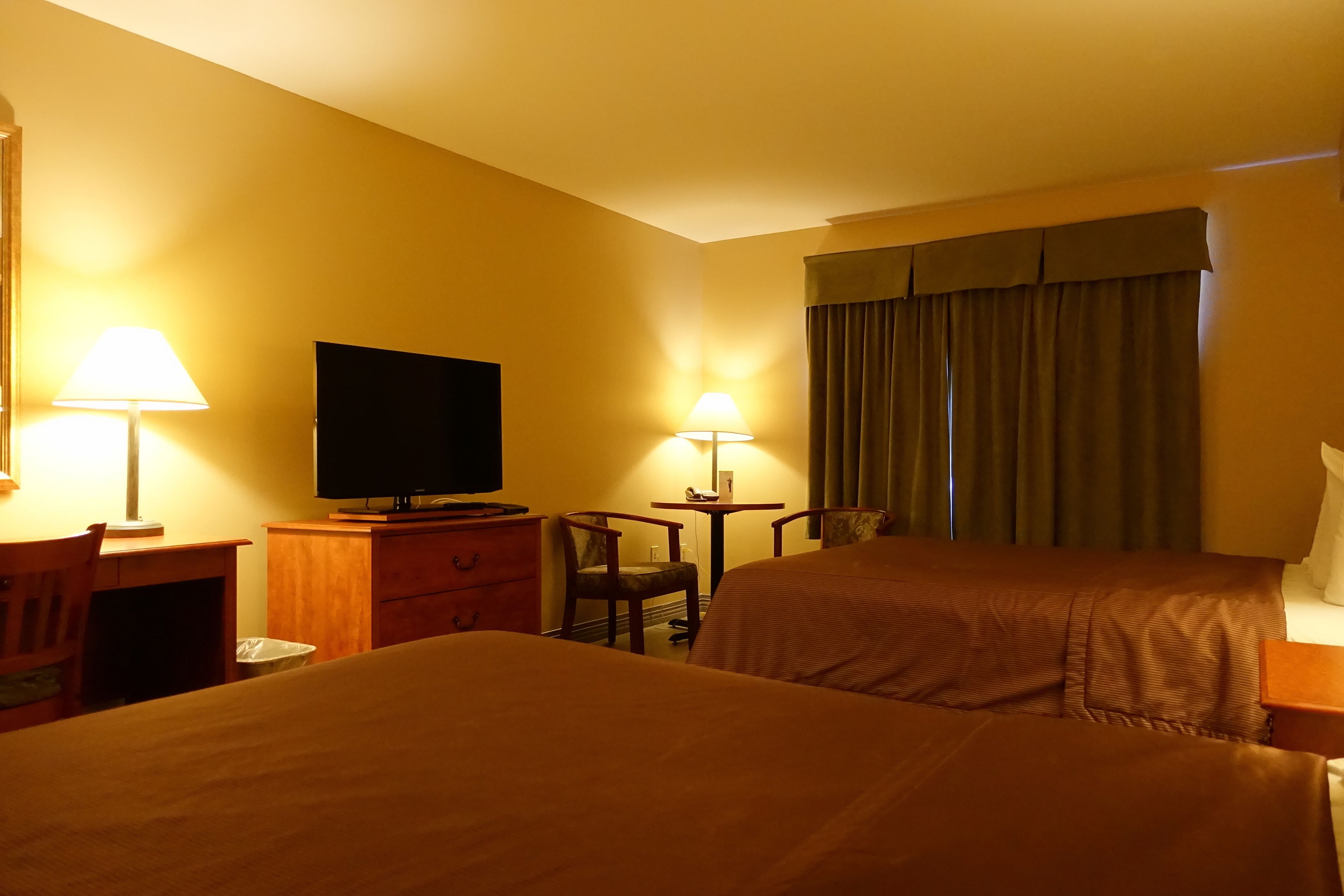 Quarto de hotel com duas camas, uma mesa com cadeira, uma televisão e duas luminárias.