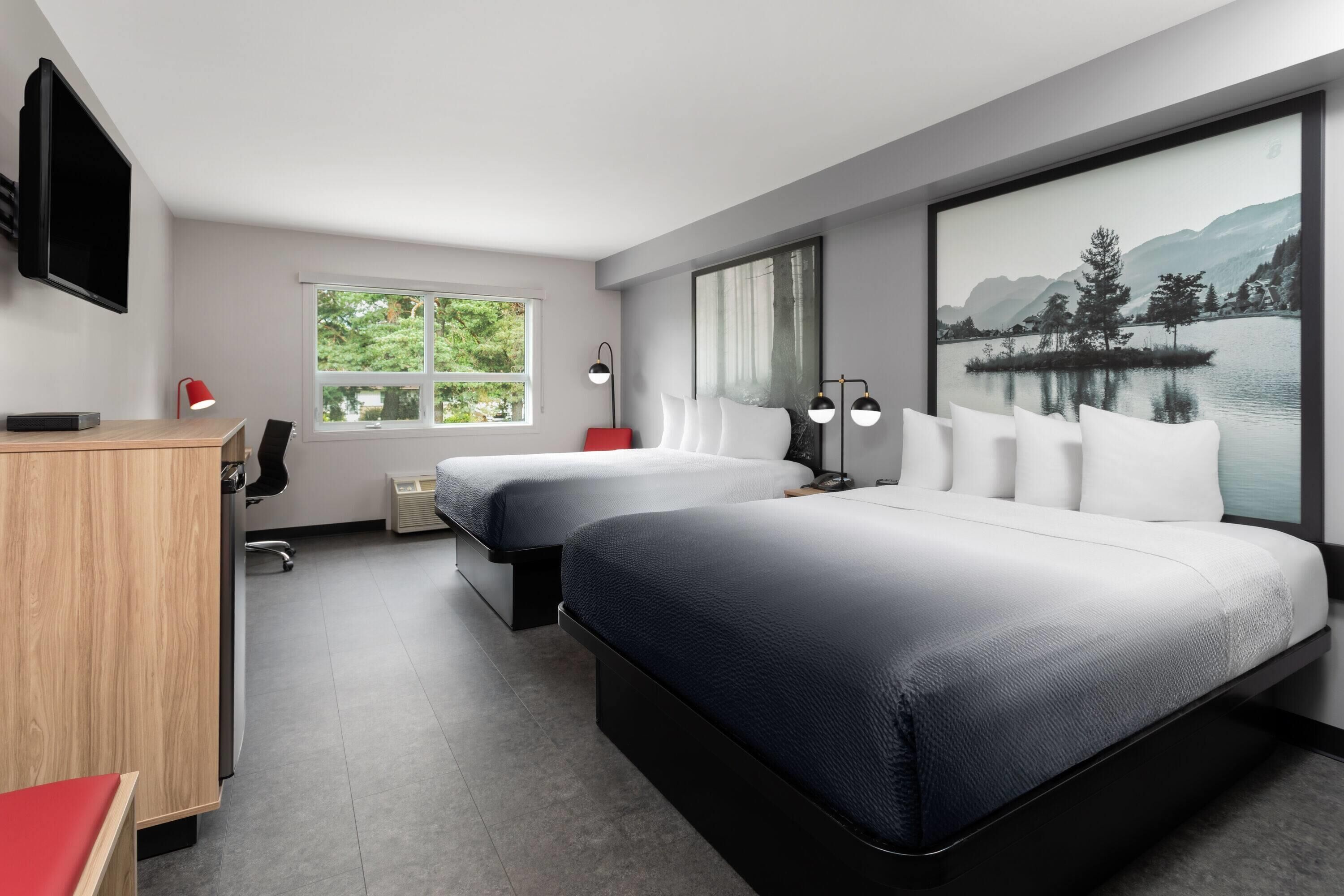 A modern hotel room with two beds, a desk, a chair, a TV, and a large window with a view of greenery.