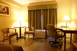 Habitación de hotel con piso de madera, dos sillones, un escritorio, una lámpara y un amplio ventanal con cortinas.