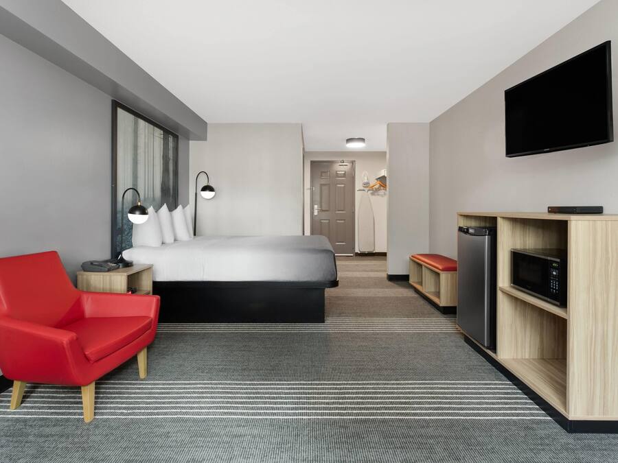 Una habitación de hotel moderna con una cama, una silla roja, un escritorio con microondas y un televisor en la pared.