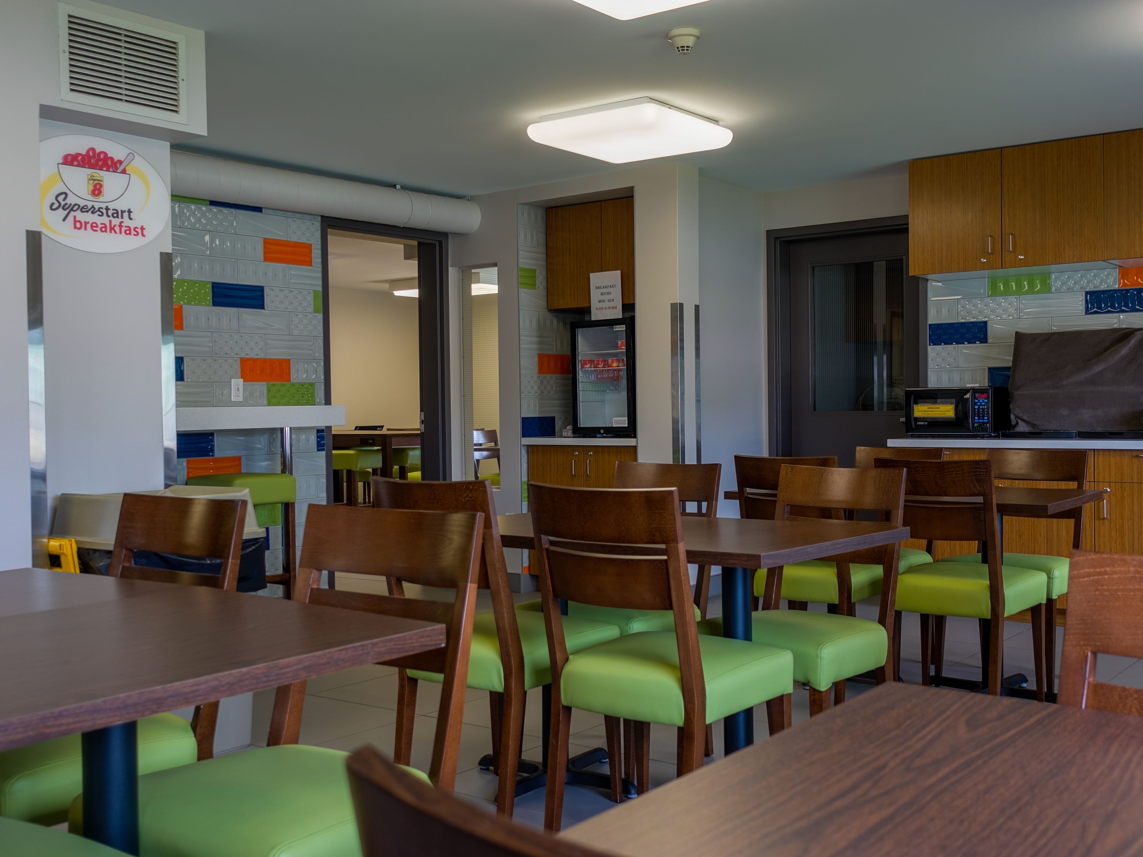 Un restaurant moderne avec des tables en bois et des chaises rembourrées de tissu vert, un four à micro-ondes et un réfrigérateur.