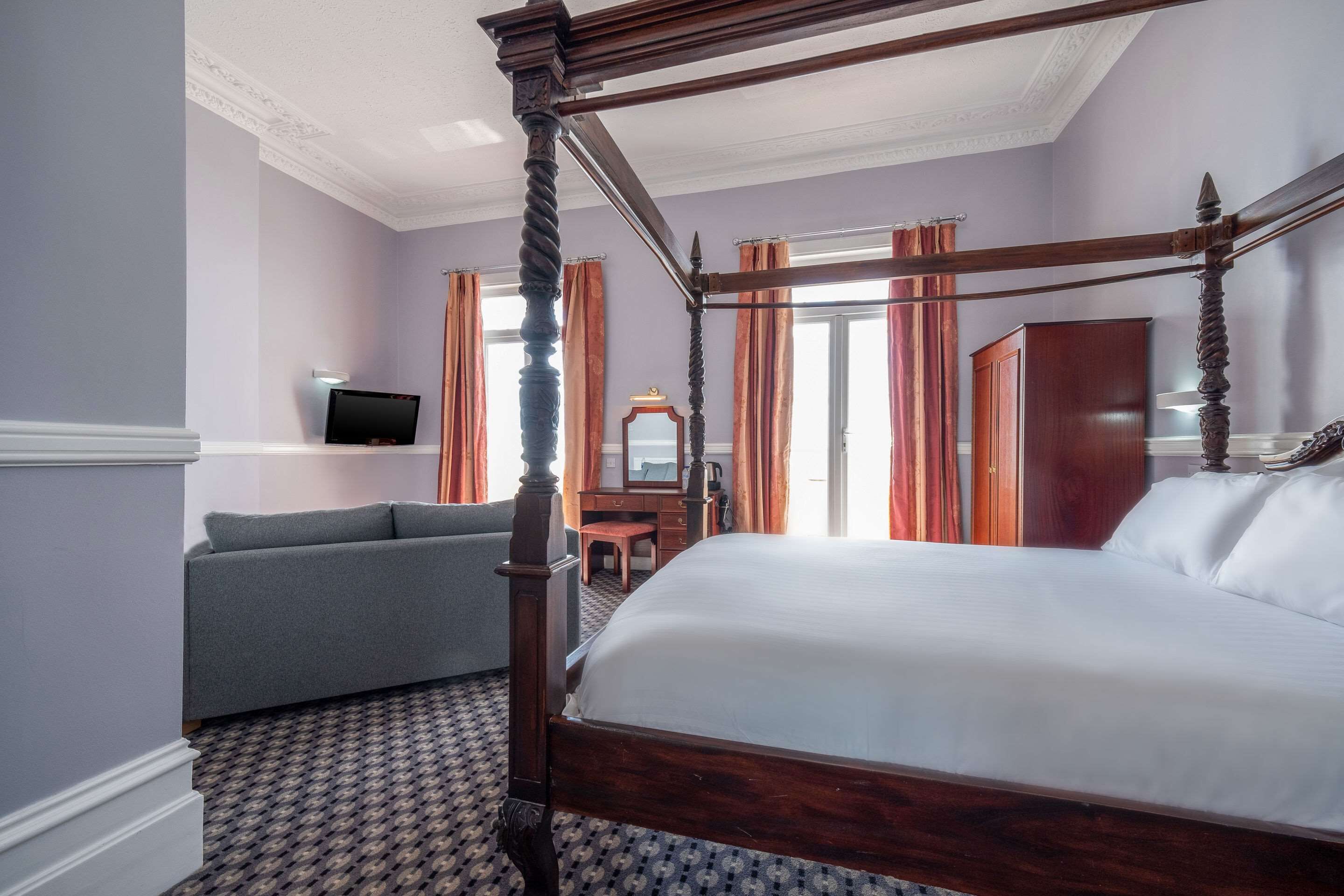 A four-poster bed with a wooden frame, a grey sofa, a small desk with a mirror, and a television on the wall.