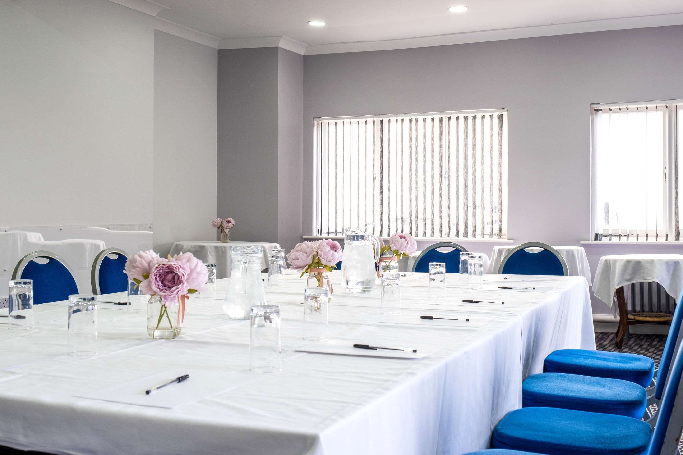 A conference room with a long table set for meetings, blue chairs, and large windows with blinds.
