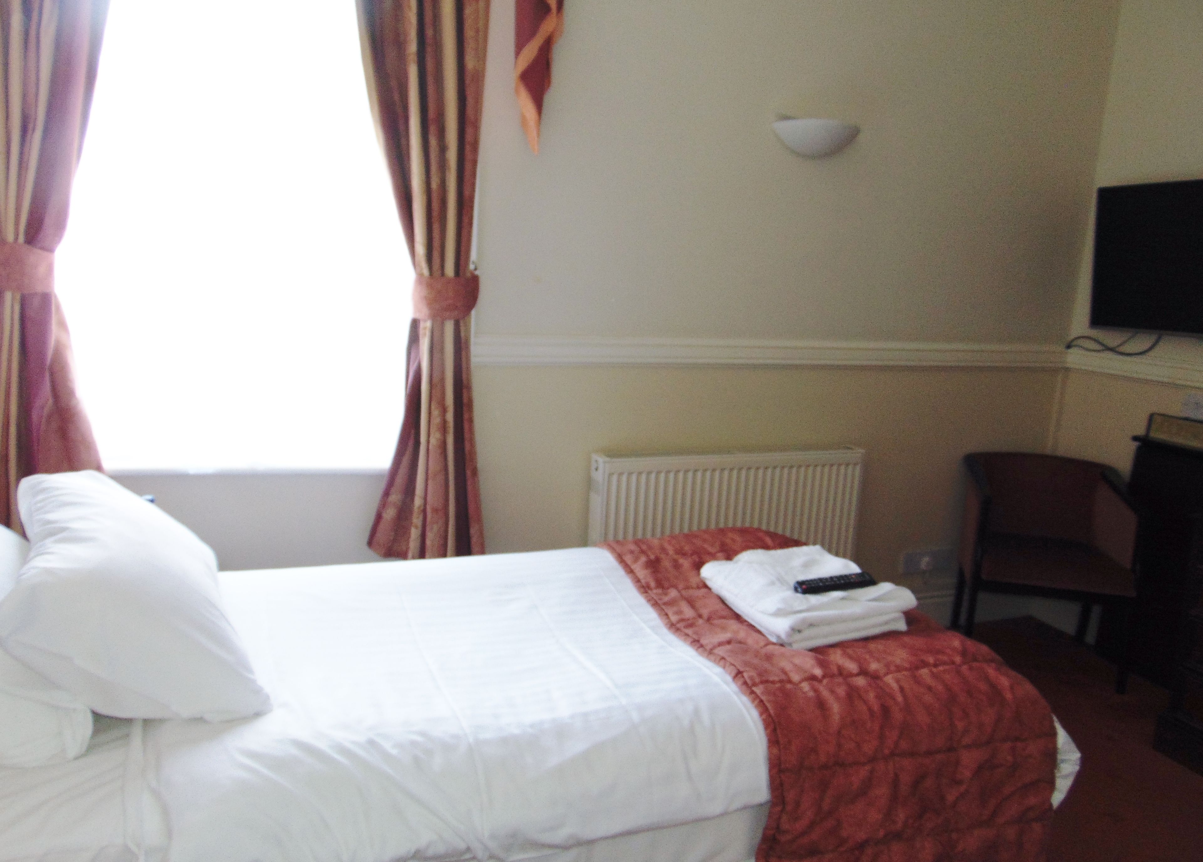Une chambre d’hôtel avec un lit, une couverture rouge, un oreiller blanc, une serviette pliée, une télécommande, une chaise et un téléviseur fixé au mur.