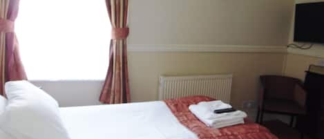 A hotel room with a bed, a red blanket, a white pillow, a folded towel, a remote control, a chair, and a television on the wall.