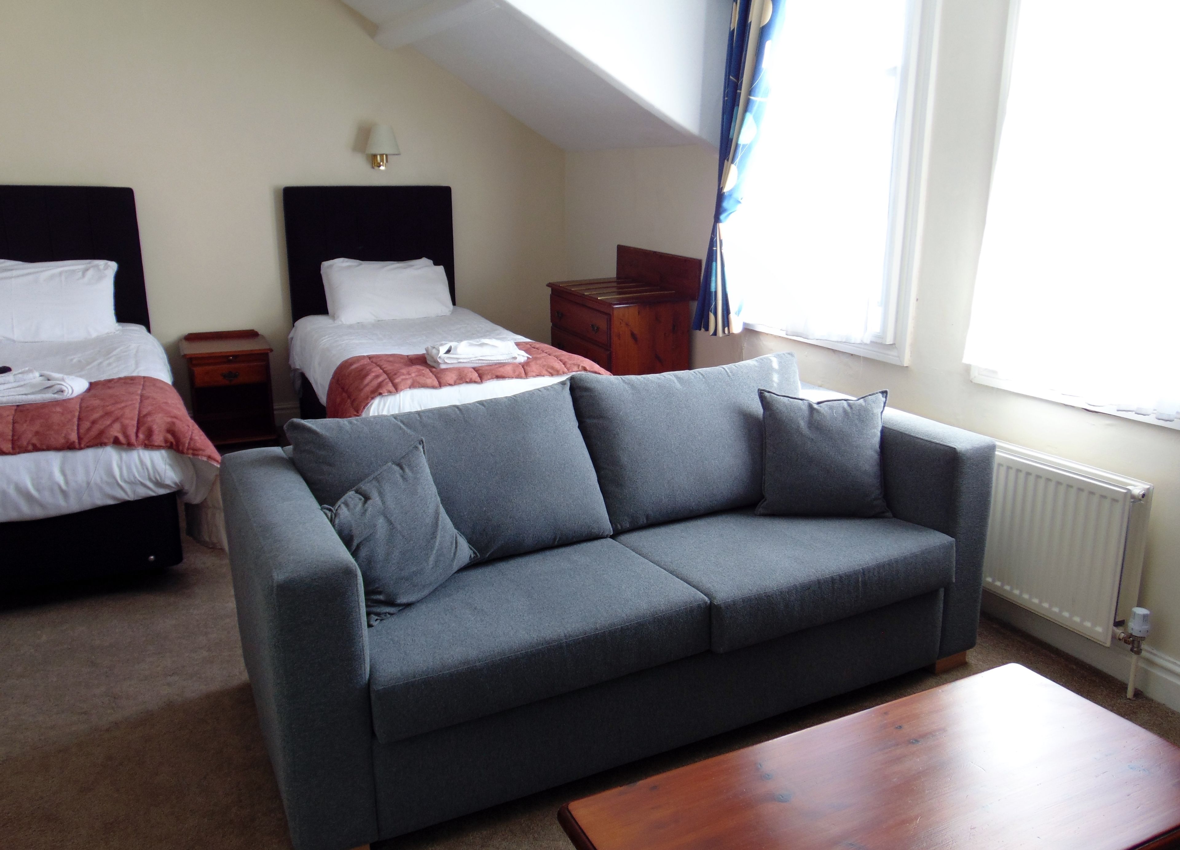 Una habitación de hotel con un sofá gris, dos camas con ropa blanca, una mesita de noche de madera, una lámpara y una ventana con cortinas.