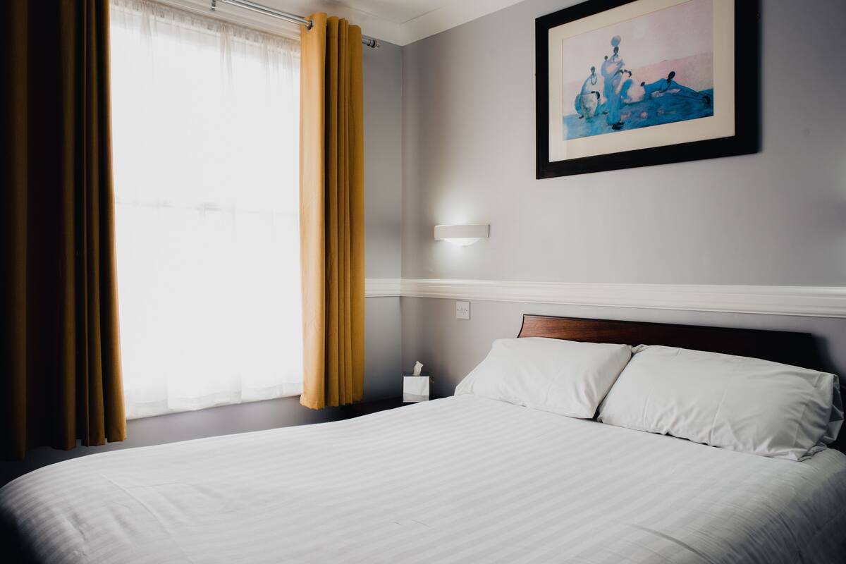 A hotel room with a large bed, a bedside table with a lamp, a painting on the wall, and a window with curtains.