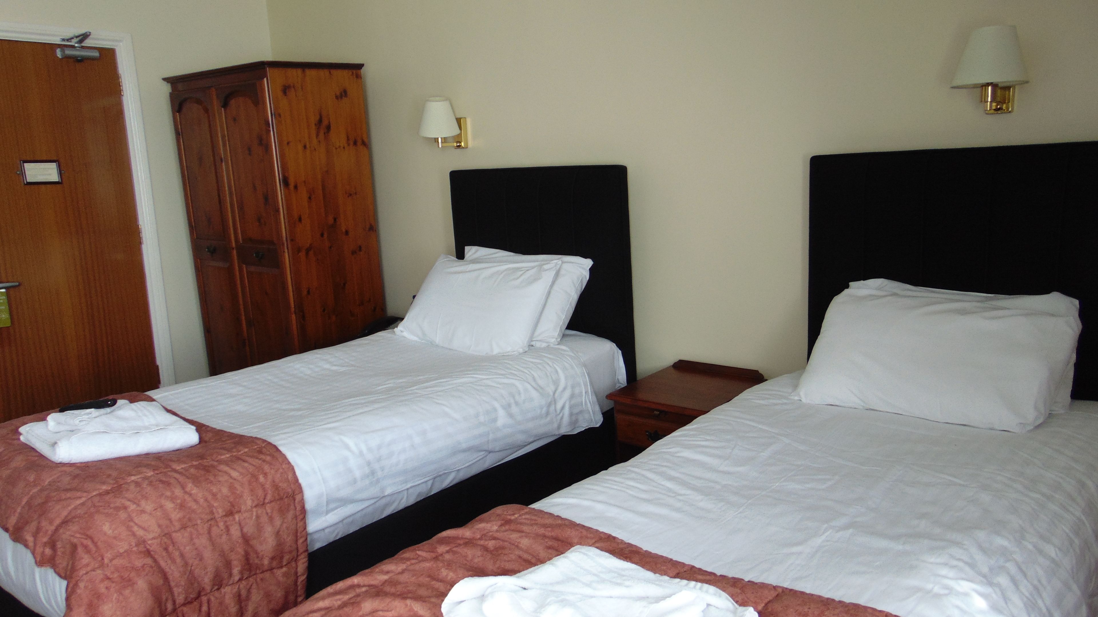 A hotel room with two single beds, a wooden wardrobe, a nightstand, and a wall-mounted lamp.