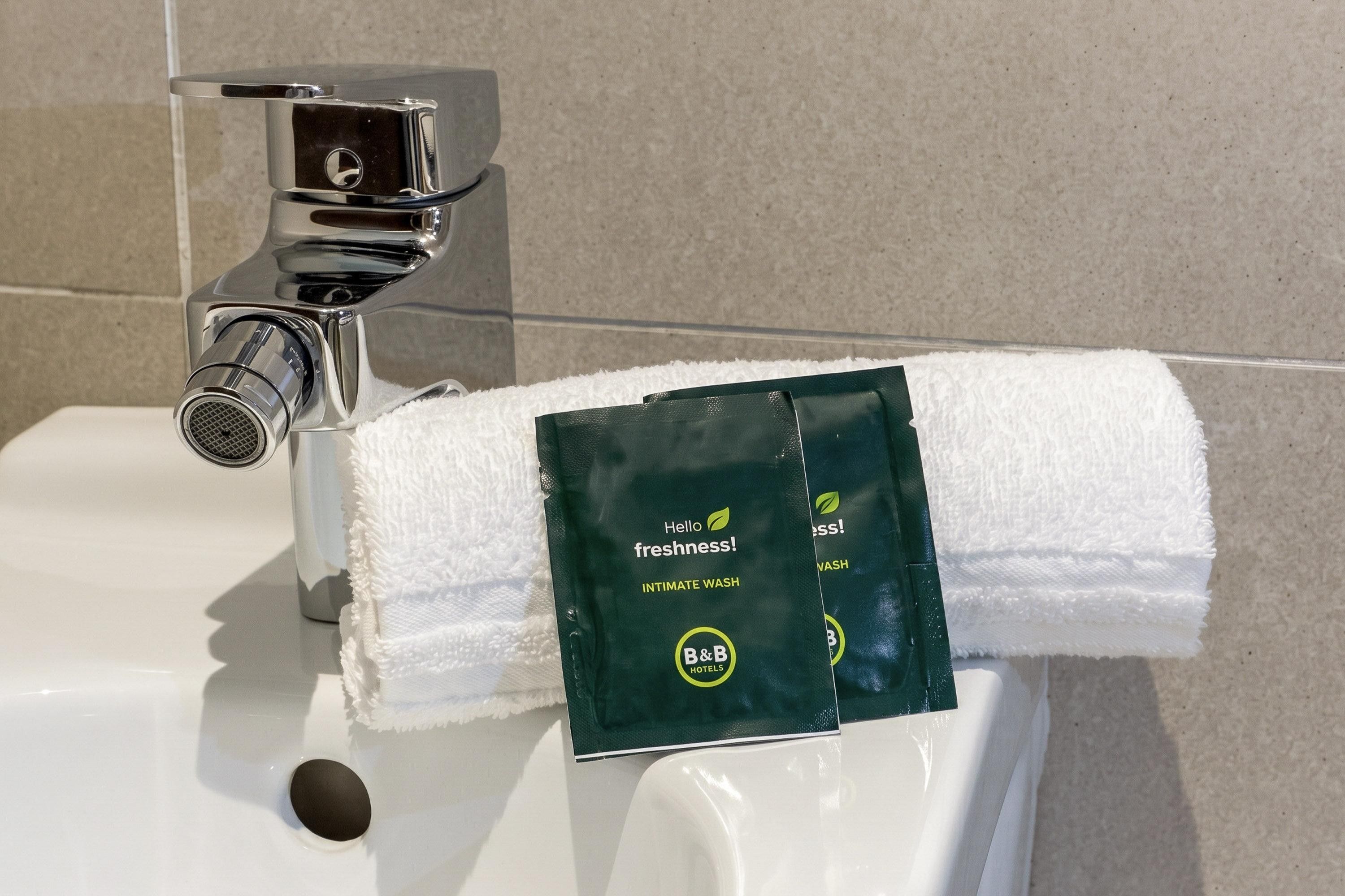 En badeværelsessink med en håndklædedispenser, der indeholder en grøn pakke mærket 'Hello Freshness! Intimate Wash' fra B&B Hotels.