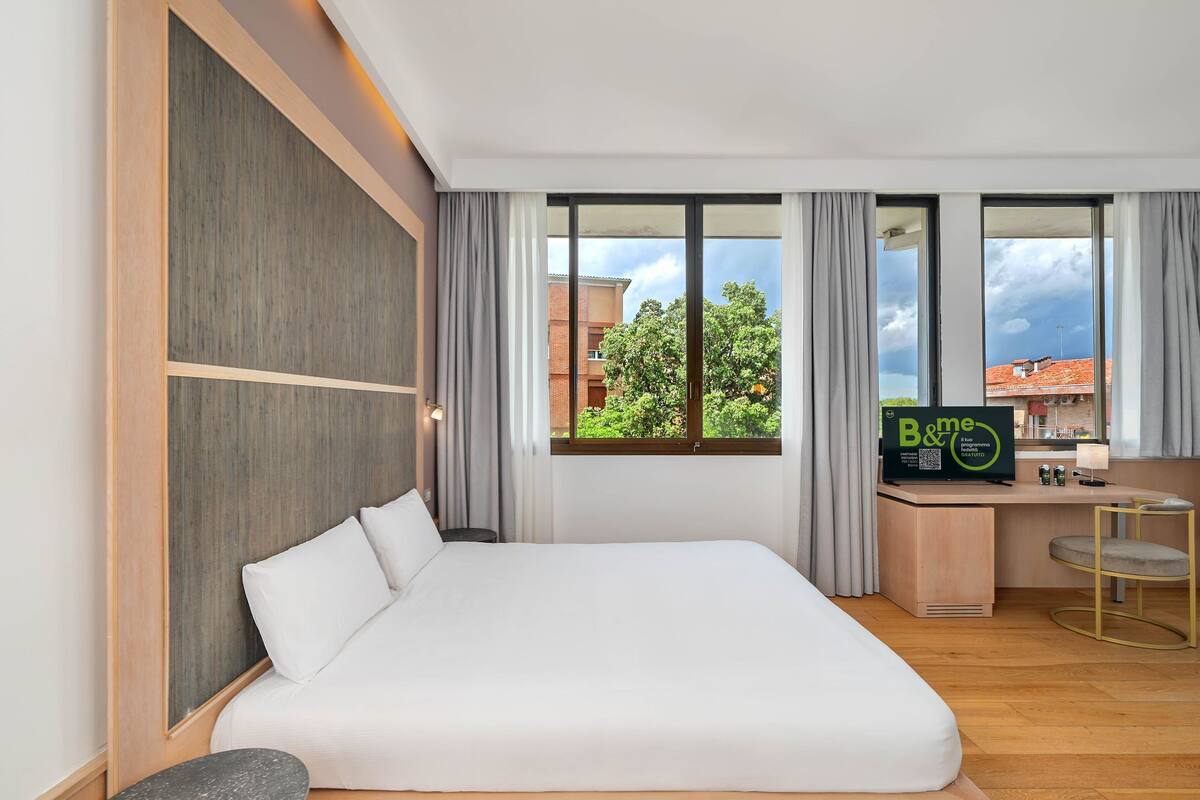 A hotel room with a bed, a desk with a computer, a chair, and a view of buildings and trees.
