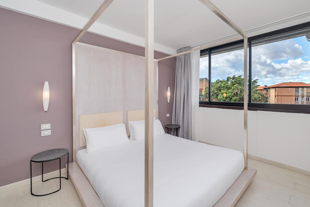 A bedroom with a four-poster bed, a small side table, a wall-mounted light, and a window with a view of buildings and trees.