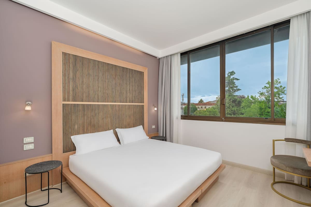 A modern hotel room with a large bed, a small side table, a chair, and a large window offering a view of greenery and buildings.
