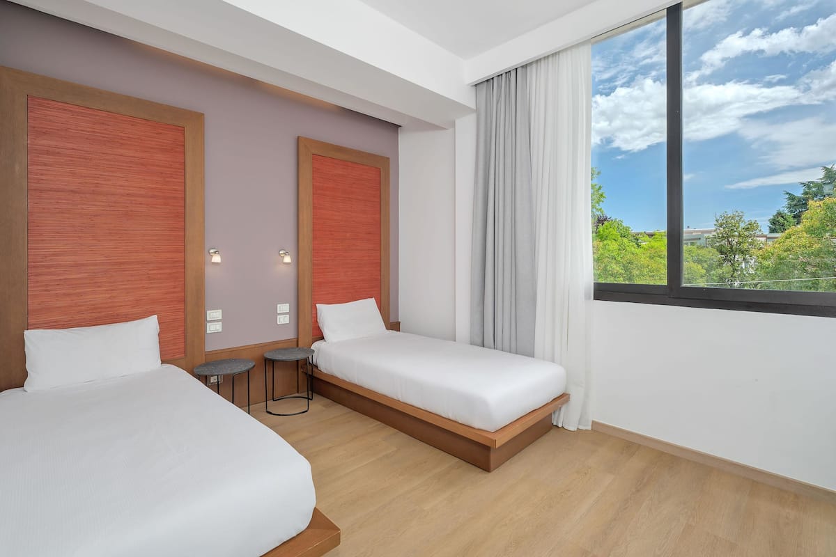 A hotel room with two beds, a small table, and a large window with a view of greenery.