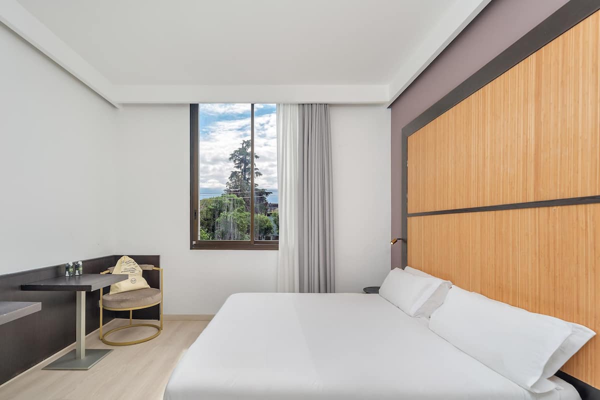 A modern hotel room with a bed, a desk, a chair, and a large window with a view of trees and a building.