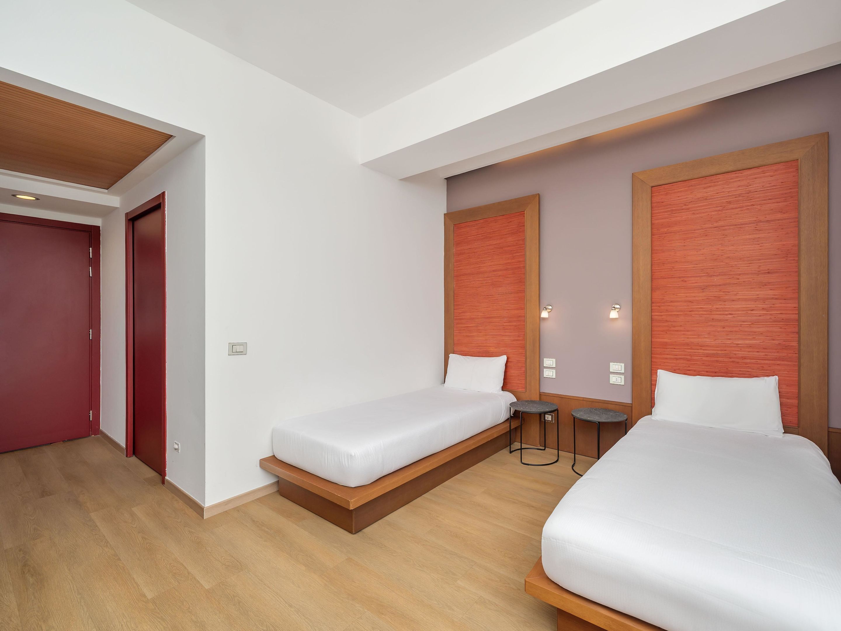 Une chambre d’hôtel avec deux lits, un plancher en bois, une porte rouge et un interrupteur sur le mur.