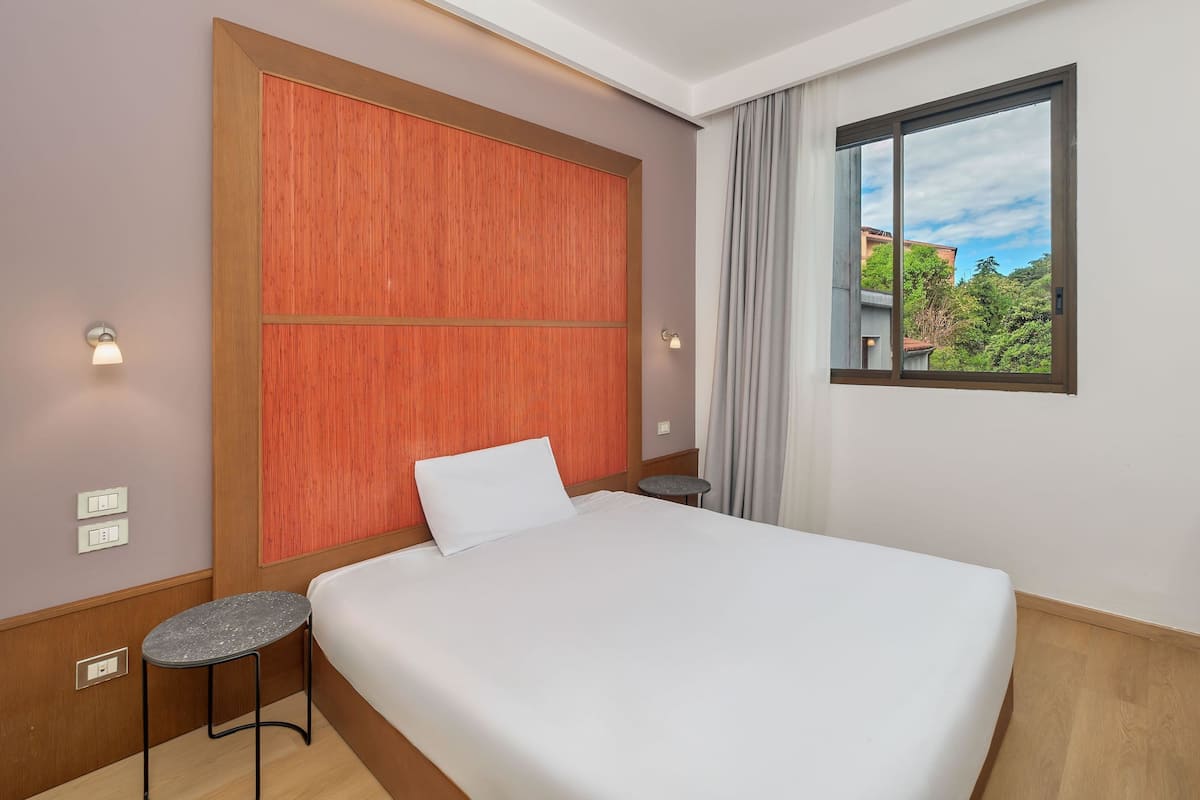 A hotel room with a bed, a small table, a nightstand, a window with a view, and wall-mounted lights.