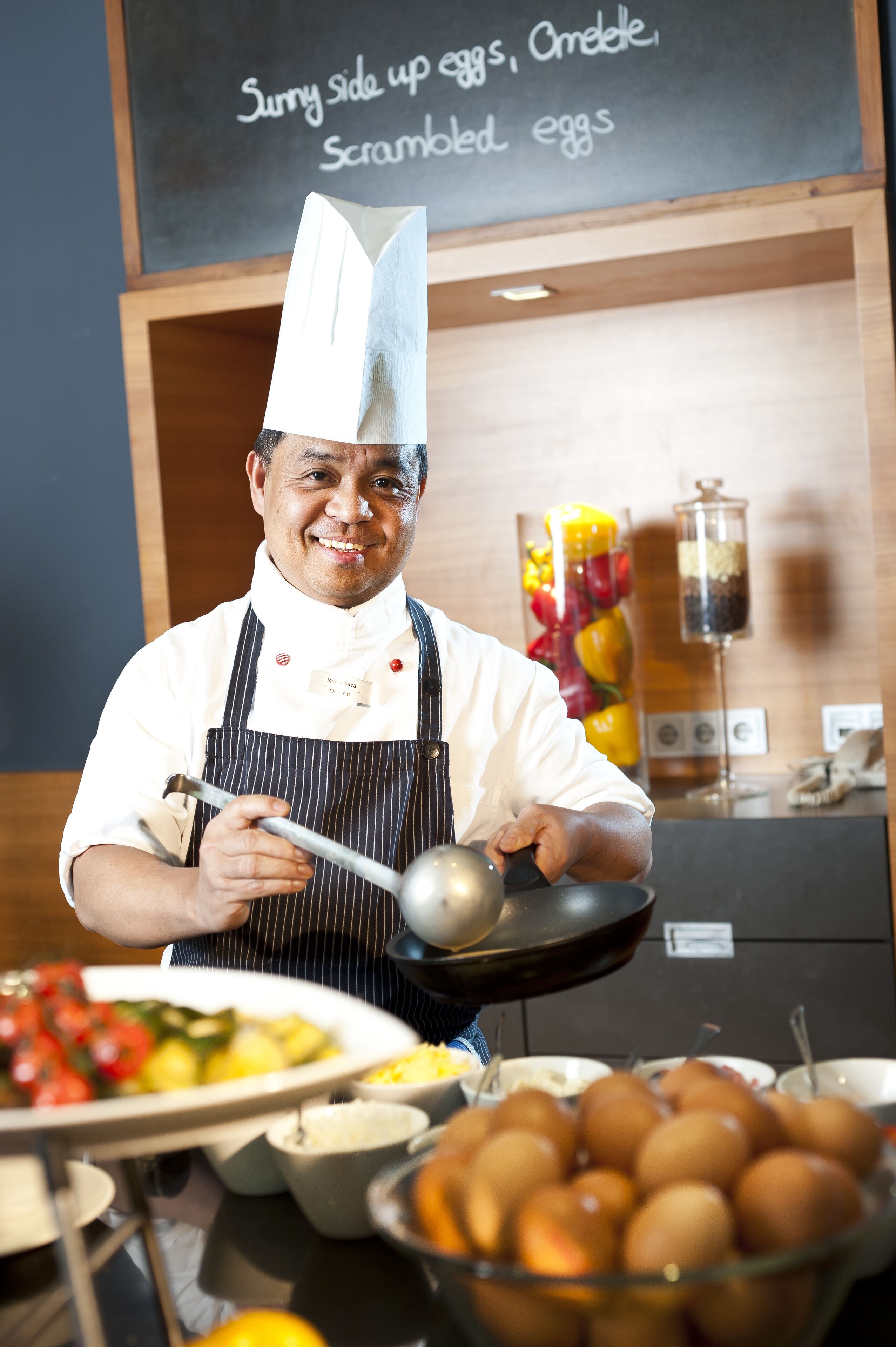 A chef in a kitchen holding a ladle, with a chalkboard menu behind him listing dishes like scrambled eggs.