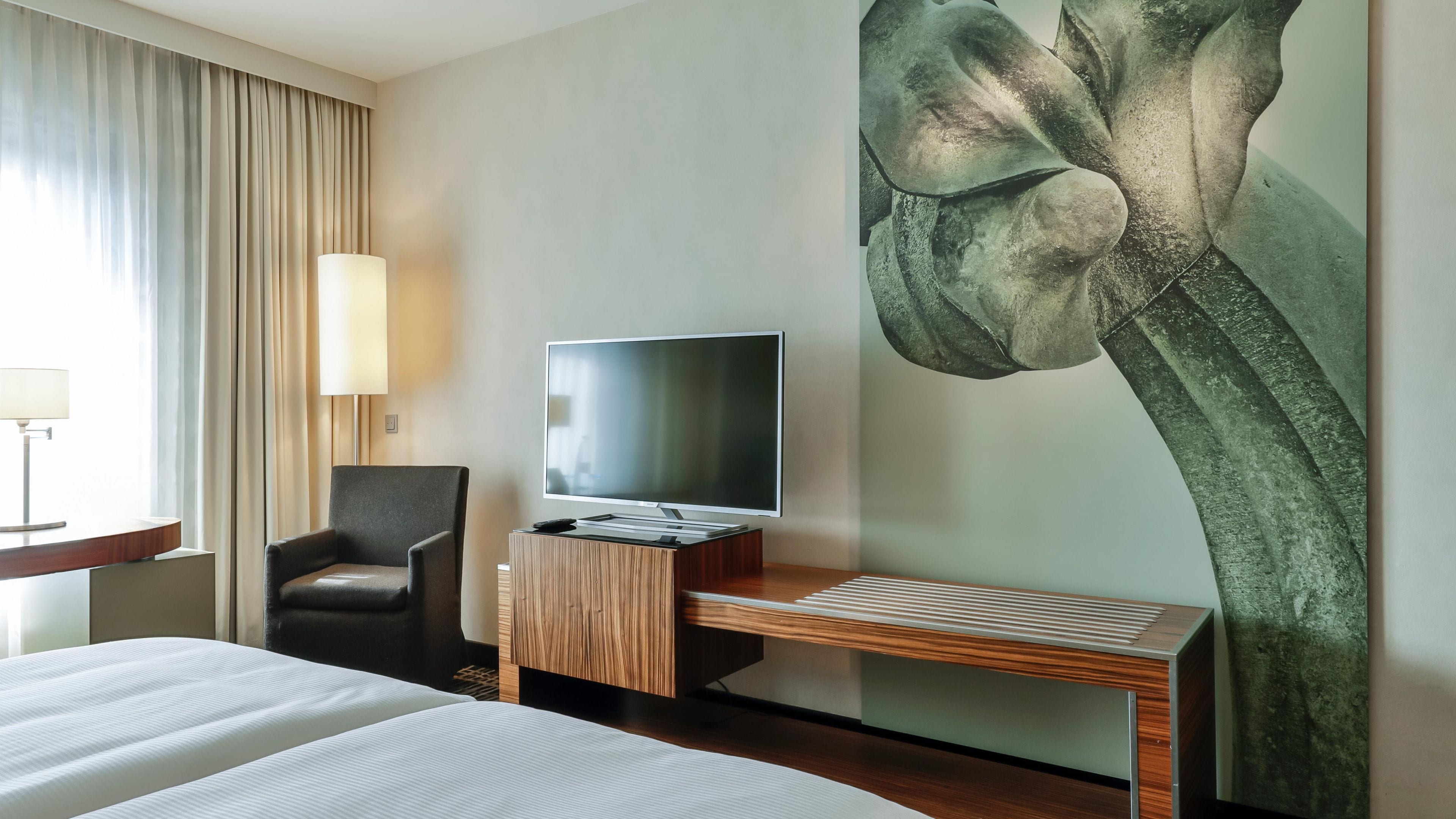 A hotel room with a bed, a desk with a flat-screen TV, a chair, and a large floral artwork on the wall.