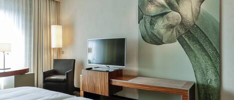 A hotel room with a bed, a desk with a flat-screen TV, a chair, and a large floral artwork on the wall.