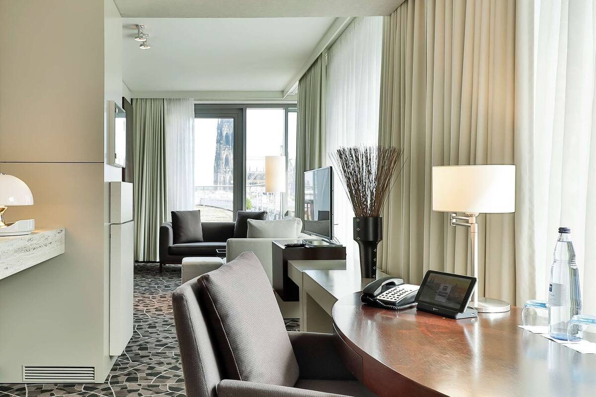 A hotel room with a desk, a chair, a sofa, a bed, and a large window with curtains.