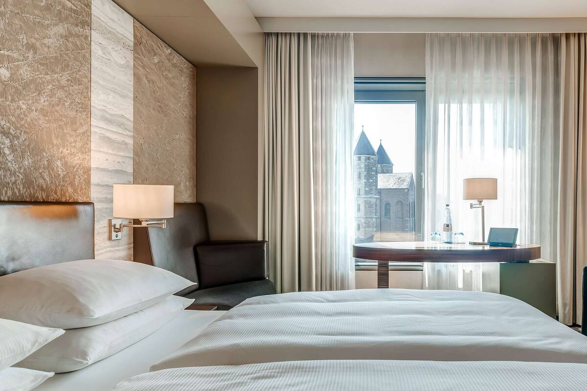 A hotel room with a large bed, two chairs, a desk with a laptop, and a view of a castle through sheer curtains.