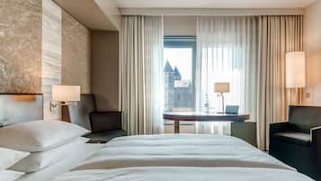 A hotel room with a large bed, two chairs, a desk with a laptop, and a view of a castle through sheer curtains.