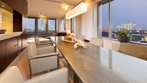 A modern hotel room with a long dining table, comfortable seating, a large window with a city view, and a decorative plant. - Dorint Hotel am Heumarkt Köln (Cologne)