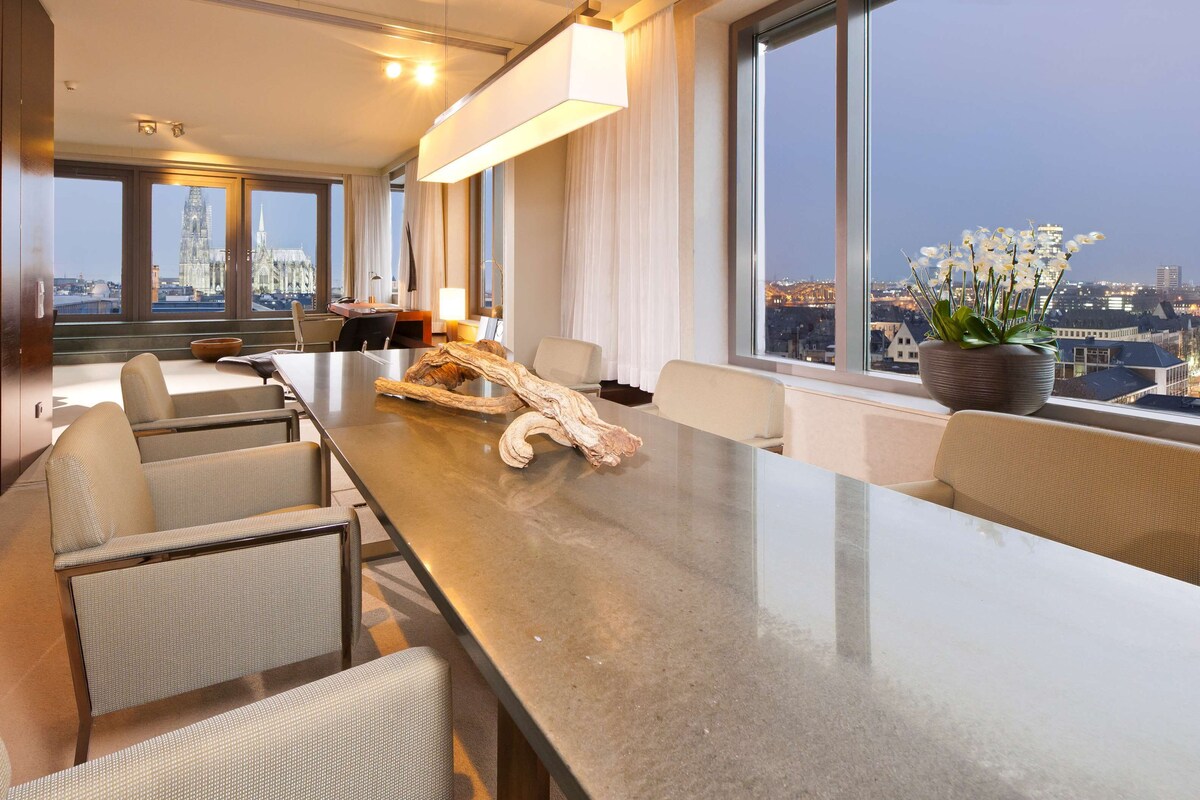 A modern hotel room with a long dining table, comfortable seating, a large window with a city view, and a decorative plant.