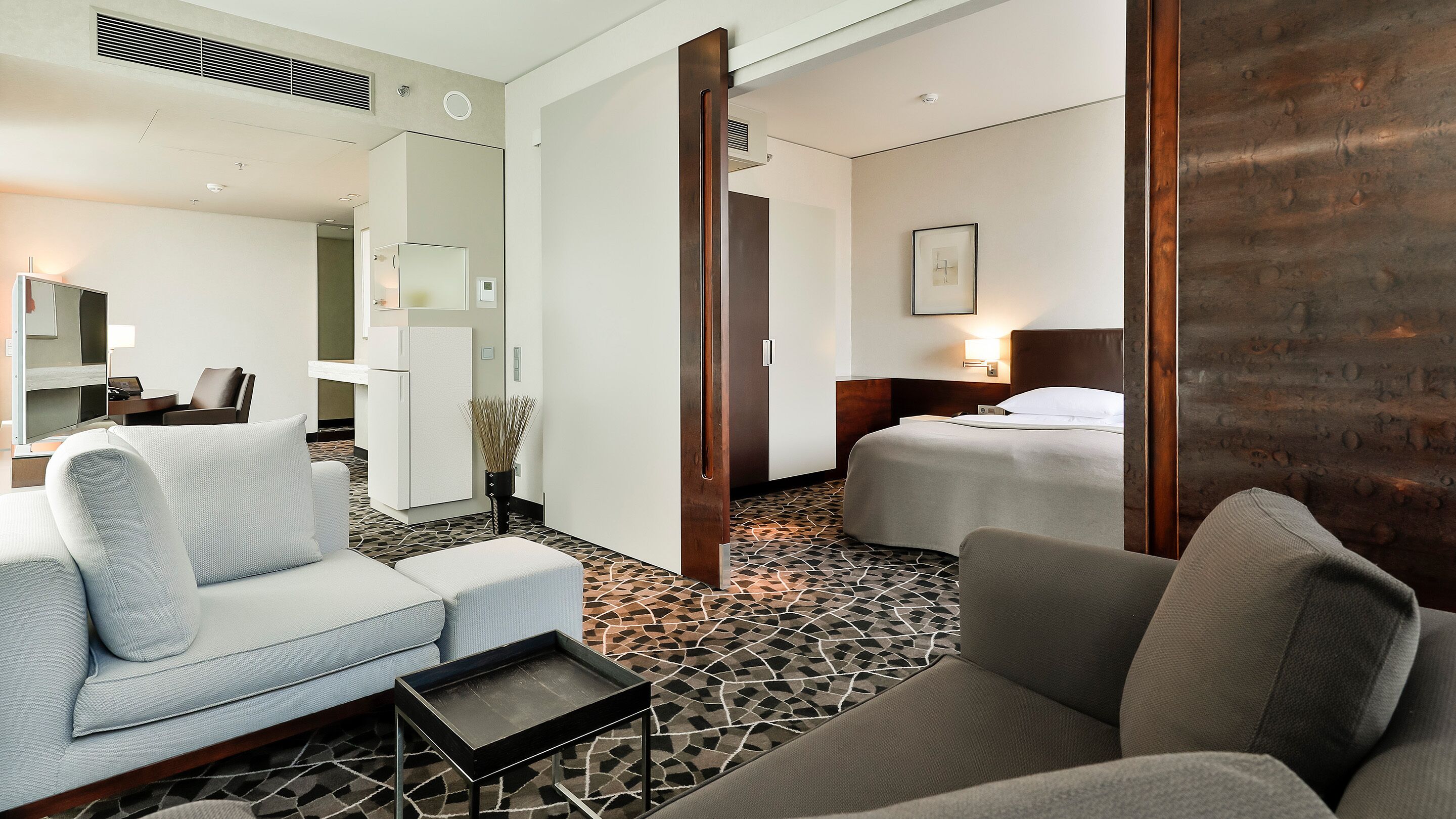 A hotel room with a sofa, a bed, a small table, a mirror, and a wall-mounted TV.